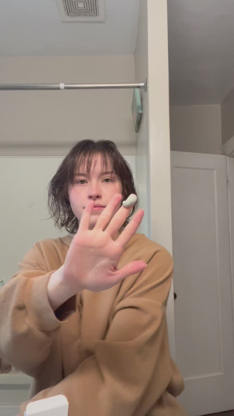 Person with short wet hair in bathroom mirror holding a fingertip of white cream toward camera