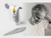 Man touching his hair beside a cosmetic tube and a smear of cream on a pale background